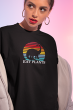 Load image into Gallery viewer, Woman wearing black vegan shirt with a picture of a dinosaur in a rainbow sun and the words eat plants.
